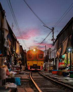 Visitors explore the bustling Maeklong Railway Market as a train approaches, surrounded by colorful stalls and local vendors. The sunset paints the sky with beautiful hues, adding to the ambiance.