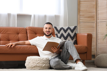 Obraz premium Young man with pouf reading magazine on carpet in living room
