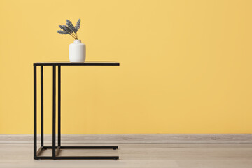 Vase with artificial flowers on table near yellow wall