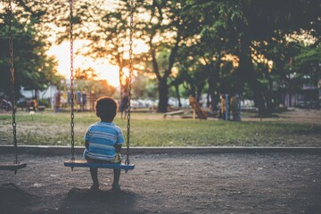 Child sits on swing in park during sunset near trees and playground in summer evening