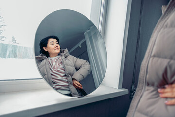young Asian woman with short black hair adjusts her coat while looking in a round mirror. Snow is visible outside the window, indicating winter time with snow
