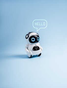 Cute futuristic robot toy with glowing eyes and speech bubble saying &ldquo;HELLO&rdquo; isolated on blue background, representing AI assistant, chatbot technology, digital communication, and virtual support.