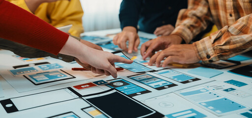A diverse group of individuals engages in collaborative brainstorming over colorful paper mockups, focusing on interface design strategies in a modern office environment. SACTR
