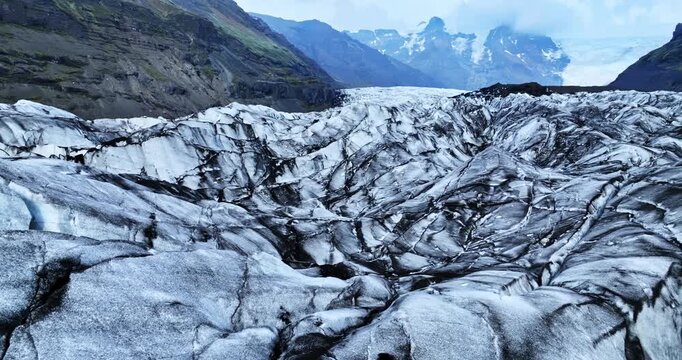 Dynamic glacier landscape with sharp ridges, deep fractures, and sweeping mountain peaks in the distance, captured from a low drone angle.