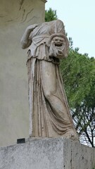 Rome, Italy - 12 January 2025. A headless statue in robes grasps a dramatic theatrical mask, standing on a pedestal beside trees and a plastered surface.