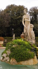 Rome, Italy - 12 January 2025. A nude female statue with worn features stands next to a jug-shaped support amid trimmed greenery and tall trees at Villa Borghese Gardens.