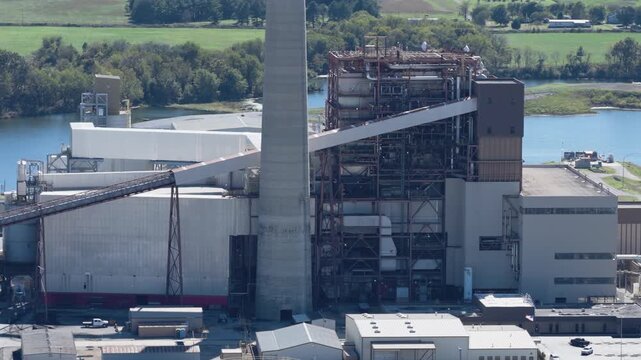 Power plant structures and surrounding infrastructure beside river and forested land, aerial medium orbit ascend reveals smokestack towering