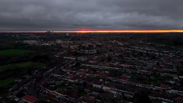 A forward-facing drone flight over Ealing in West London at dusk. The drone passes above rooftops and houses beneath thick cloud cover, with a striking horizontal orange glow from the setting sun.