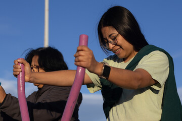 Young woman smiling and exercising on an outdoor elliptical machine with a friend, enjoying a healthy lifestyle and fitness together in a park