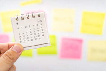 Hand holding January 2026 mini calendar with blurred colorful sticky notes background. new year planning, goal setting, organization, and a fresh start in a busy work or personal life environment.
