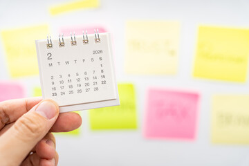 Hand holding February 2026 mini calendar with blurred colorful sticky notes for planning concept. project tracking, monthly goal setting, and time management in a creative workspace.