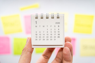 Hand holding January 2026 mini calendar with blurred colorful sticky notes background. new year planning, goal setting, organization, and a fresh start in a busy work or personal life environment.
