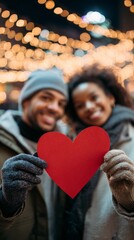 Black couple holding red heart paper symbol at night with holiday lights backdrop, during Valentine's day, vertical photo
