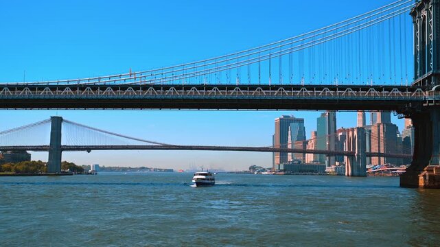 Ferry crossing the East River framed between the Manhattan and Brooklyn bridges. The view from the water shows the lower decks of two iconic suspension bridges with the downtown Manhattan skyline 