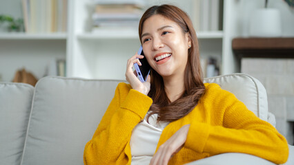 Happy young asian woman using mobile apps in living room,Smiling young asian female using smartphone sitting on couch at home.Messaging,Online Shopping,celebrate success,chatting online,Social Media