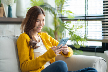 Happy young asian woman using mobile apps in living room,Smiling young asian female using smartphone sitting on couch at home.Messaging,Online Shopping,celebrate success,chatting online,Social Media