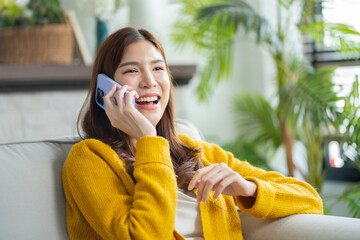Happy young asian woman using mobile apps in living room,Smiling young asian female using smartphone sitting on couch at home.Messaging,Online Shopping,celebrate success,chatting online,Social Media