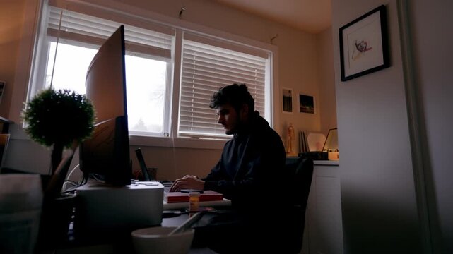 Low angle static shot of a man working at a computer in a home office, with window blinds closed and warm indoor lighting