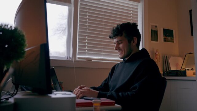 Static side view of a man typing on a keyboard while working at a computer in a home office with soft window light and warm interior lighting