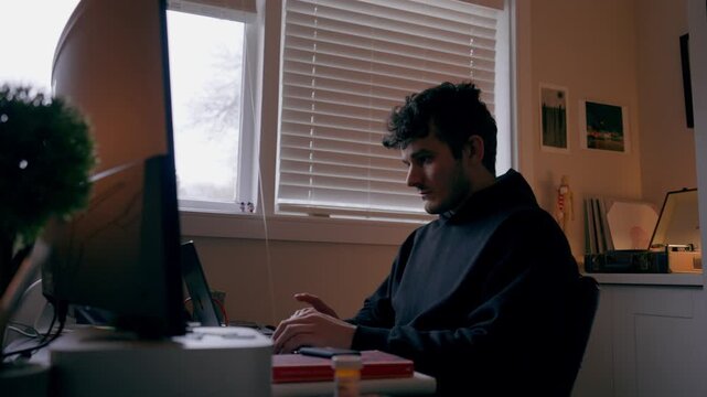 Close-up shot of a focused man working at a computer in a home office, with soft window light and warm indoor lighting