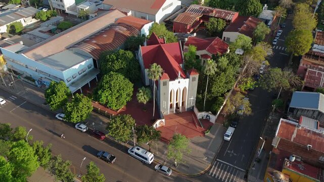 Overhead drone orbit with a camera tilt down captures the red-roofed Parroquia Inmaculada Concepci&oacute;n church nestled among green trees and city streets in Posadas, Misiones, Argentina.