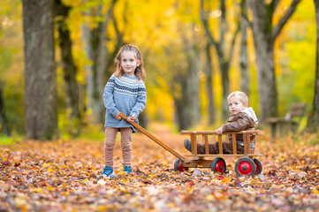 Gl&uuml;ckliche Geschwister beim Spielen mit einem antiken Leiterwagen in einer goldenen Herbstallee
