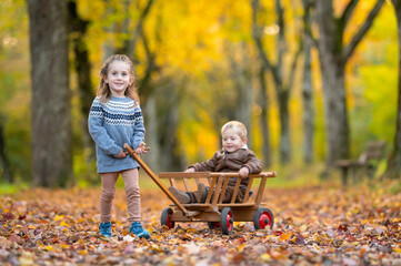 Gl&uuml;ckliche Geschwister beim Spielen mit einem antiken Leiterwagen in einer goldenen Herbstallee
