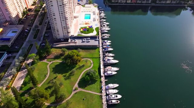 Top-down aerial drone view of a South Florida waterfront. The marina is lined with luxury boats, calm green waters, a landscaped park with walking paths, and adjacent high-rise condominium buildings.