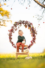 Lachendes Kleinkind sitzt auf Holzleiter in herbstlicher Natur mit K&uuml;rbissen und Bl&auml;tterkranz
