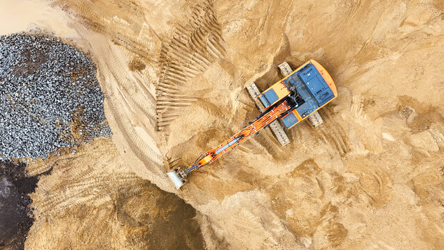 A large orange excavator is sitting in the sand, surrounded by rocks and dirt. The scene is desolate and barren, with no signs of life or vegetation. The excavator is the only object in the image