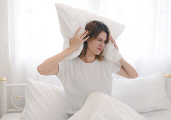 Obraz premium A young woman, disturbed by the noise from her neighbors, covers her ears with a pillow while trying to sleep in her bed at home in the early morning.