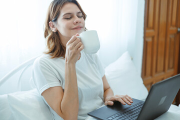 A young woman is enjoying a relaxing morning in bed, sipping coffee before starting work on her laptop. A bright morning, the idea of ​​working from home.