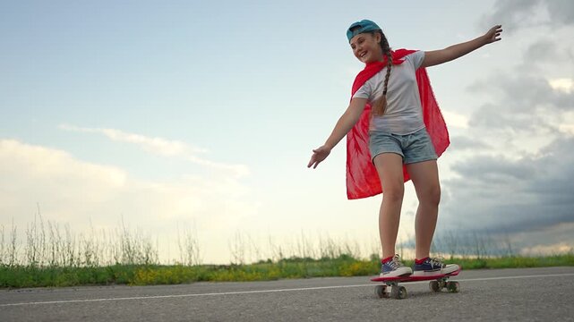 Girl rides skateboard with superhero cape on road. Child plays hero while skating outdoors. Kid with cape balances on skateboard. Girl superhero enjoys skating. Road play with child hero cape.