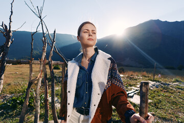 Naklejka premium Woman portrait in mountains countryside with sunlight on her face, rural fence and travel vibe. Confident traveler in denim shirt and cozy jacket exploring scenic field and valley.