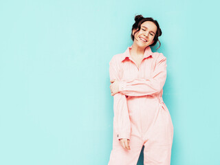 Obraz premium Young beautiful smiling female in trendy summer pink overalls. Sexy carefree woman posing near blue wall in studio with two horns. Positive model having fun and going crazy. Cheerful and happy