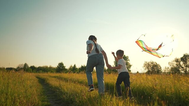 Mother and son fly rainbow kite running in meadow field. Woman with boy plays flying kite at sunset. Parent child run holding hands outdoors. Mother and son with kite in meadow at sunset.