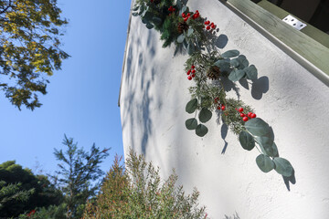 Festive eucalyptus garland with red berry decoration hanging on white exterior wall during sunny day bringing joyful holiday spirit to outdoor home design © Core