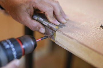 Concentrated male carpenter hand carefully installing metal hinge onto wooden plank using power drill in workshop environment showing precise craftsmanship and manual labor skill with sawdust