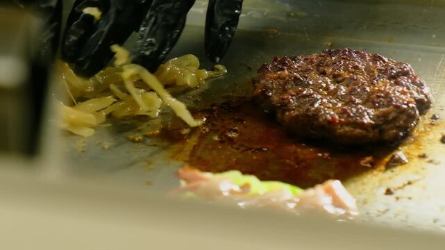 Cook hands put pickled onion near frying cutlet on grill closeup. Professional chef roasts burger ingredients in restaurant kitchen