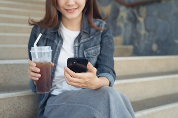 Happy young asian woman sitting on the steps holding a cup coffee, using mobile smart phone to browsing social network, smiling joyfully, modern people lifestyle