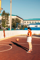 Asian teen girl in red t shirt captured mid-throw as she aims a basketball at the hoop on an outdoor court. the dynamic upward angle emphasizes the ball's motion, motion blur