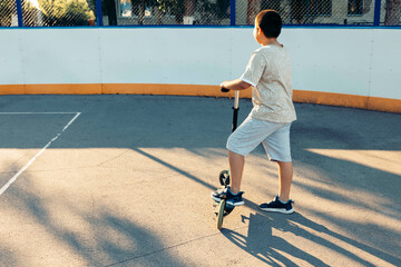 Unrecognizable boy riding scooter on outdoor court, wearing beige shirt and gray shorts, focused expression, evening sun casting long shadows, fenced area surrounded by trees