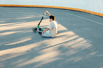 A young Caucasian male teenager sits on the ground, holding a green scooter. The scene is set on a concrete surface with shadows from nearby structures.