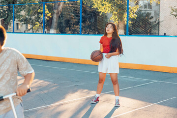 A young Hispanic girl with long brown hair holds a basketball on a court. A boy rides a scooter in the background. The scene is bright and sunny.