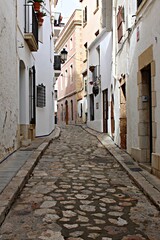 Fototapeta premium Narrow medieval street with cobblestone pavement in Sitges, traditional Spanish old town architecture, white houses with balconies. Historic European alley, travel destination and charming village 