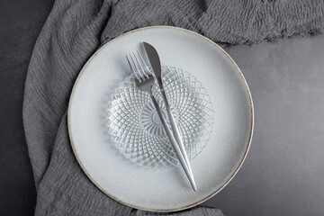 Horizontal top view of light ceramic plate with glass plate and metal cutlery on dark gray fabric. Copy space. Concept of minimalist table background