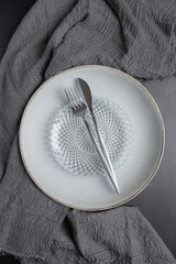 Vertical top view of light ceramic plate with glass plate and steel cutlery on gray fabric. Minimalist and modern table composition for dining, decor and hospitality. Concept of modern table styling