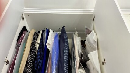 An open white closet reveals neatly hung clothes in varied colors and fabrics, some protected with plastic covers. The organized arrangement highlights tidy storage and home organization