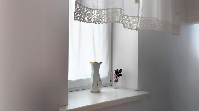 A white windowsill holds a tall vase as sheer curtains sway softly in natural light. The gentle movement creates a calm, serene atmosphere with a minimalist and peaceful touch