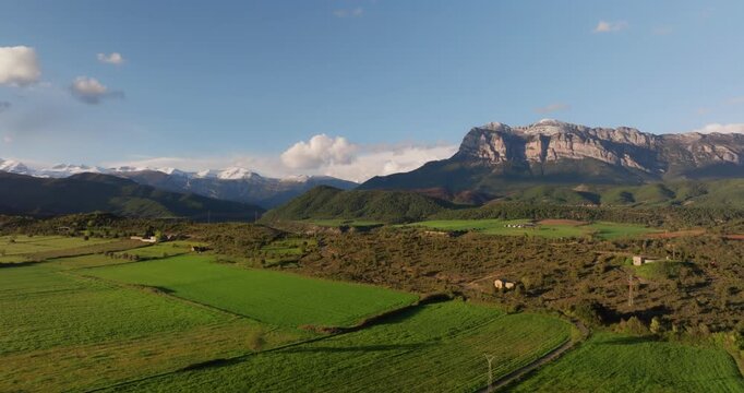 Drone ascends over the lush green valleys of A&iacute;nsa, Spain, revealing the dramatic limestone face of Pe&ntilde;a Monta&ntilde;esa against the snow-capped Central Pyrenees.